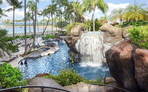 Zona de piscina en un resort con una cascada, rodeada de palmeras y sillas de descanso.