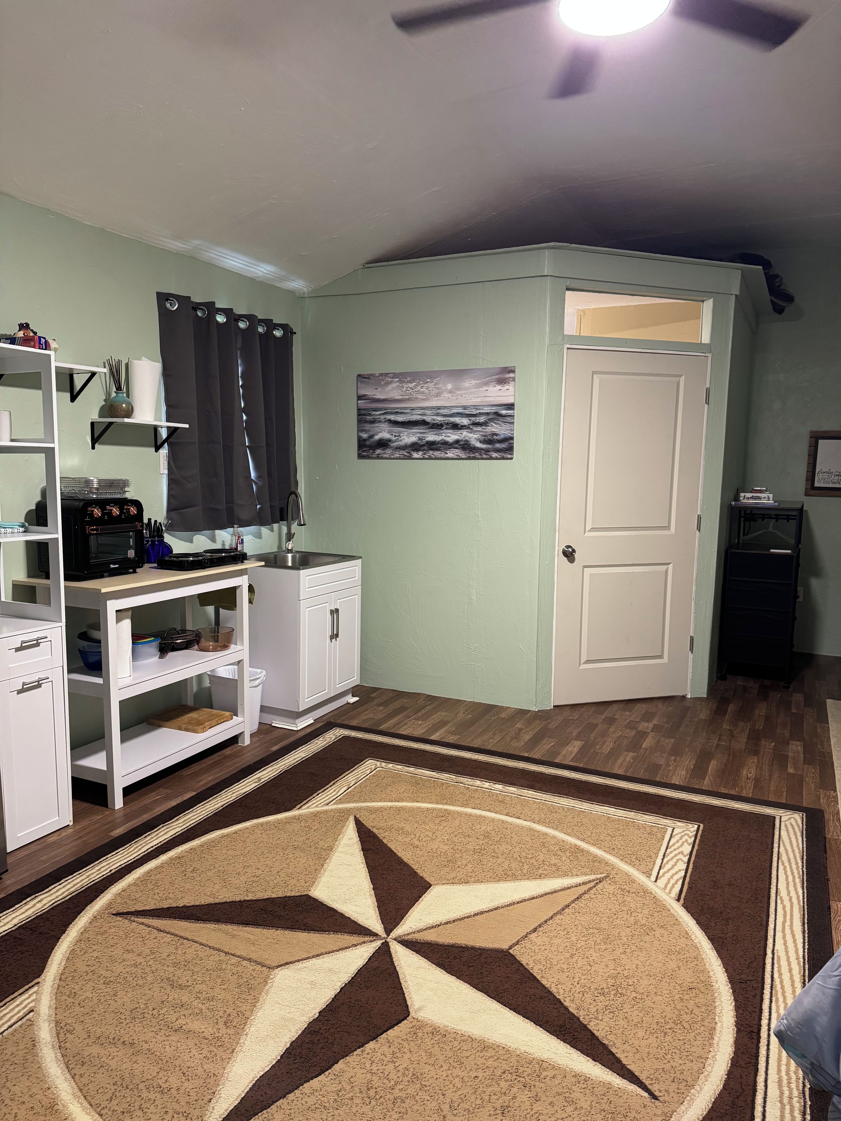 A room with a kitchen area, a white door, a painting of a seascape, and a patterned rug.