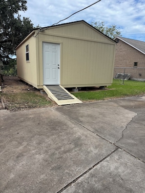 A small, single-story shed with a door and a small window, situated next to a brick building.