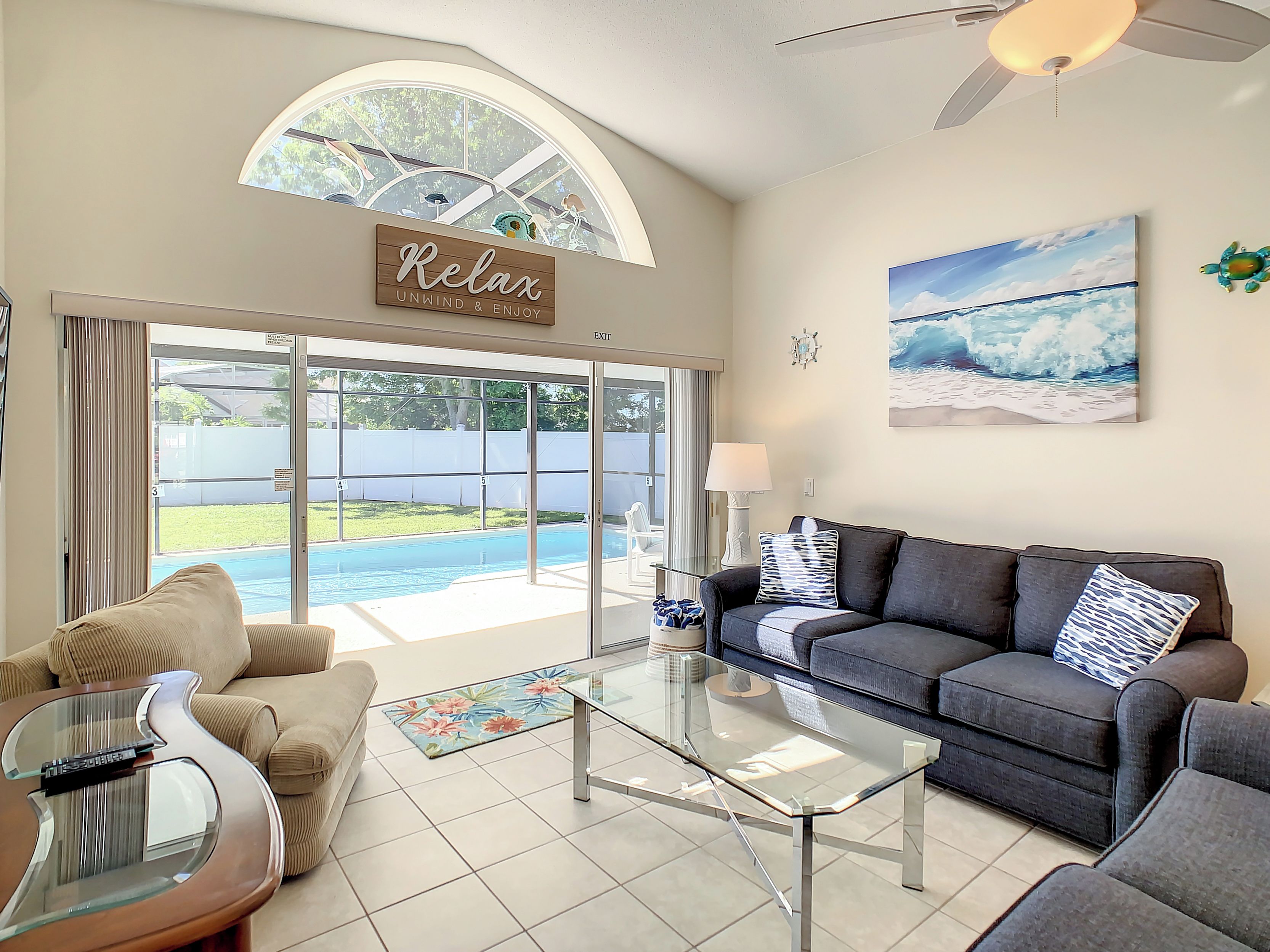 A living room with a glass coffee table, a sofa, a couch, and a piano. There is a large window with a view of a swimming pool and a sign that says "Relax."