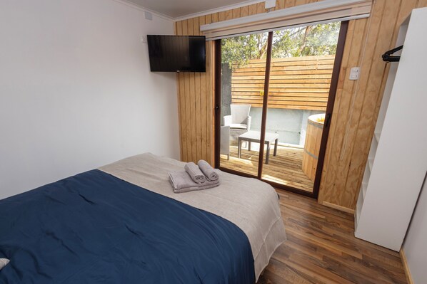 Une chambre avec un lit, un téléviseur fixé au mur et une porte-fenêtre coulissante donnant sur un espace extérieur doté d’une terrasse en bois.