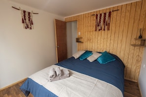 A bedroom with a wooden panel wall, a bed with blue bedding and teal pillows, a wooden door, and a small shelf with a candle. - New Listing (Valdivia)