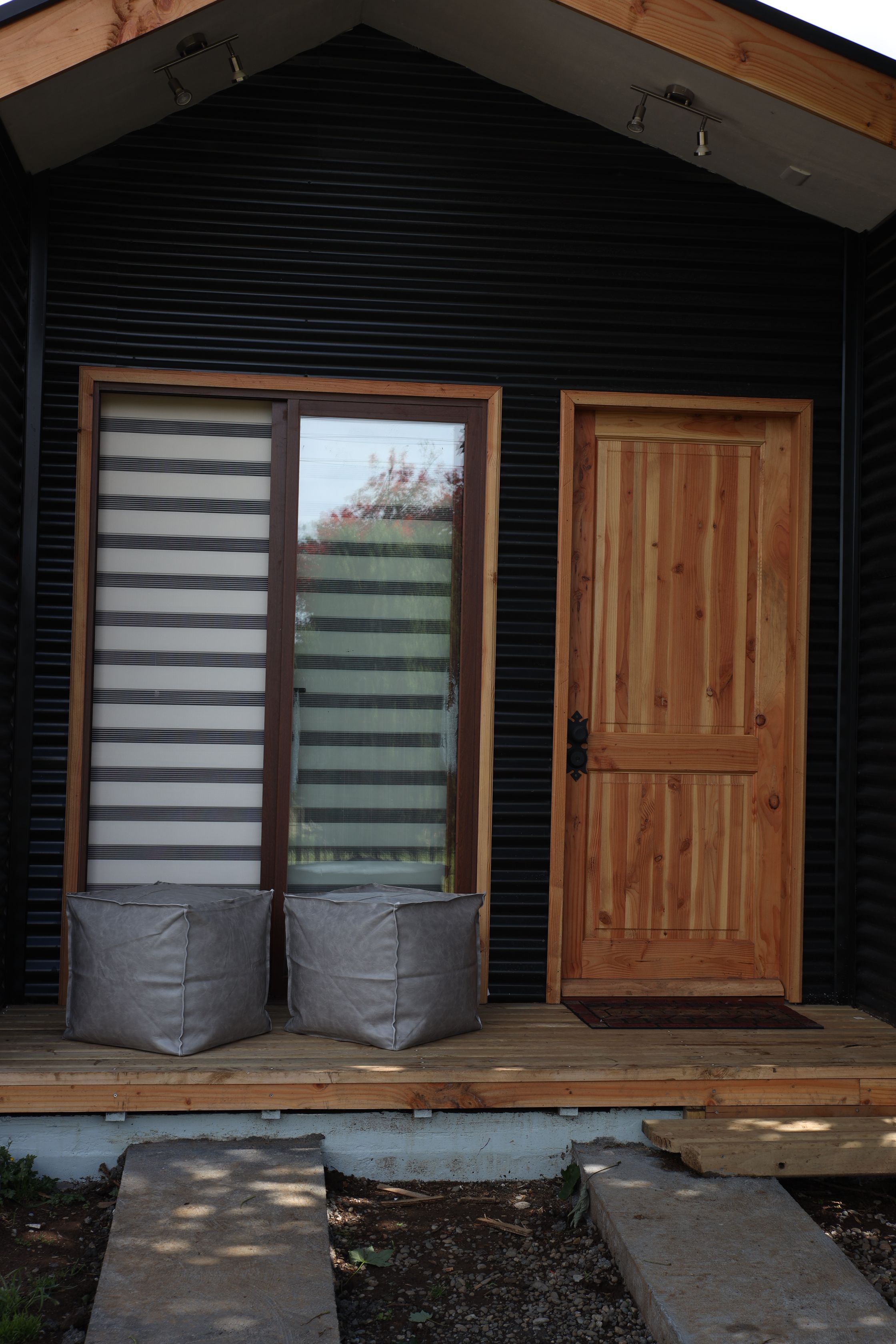 Une porte en bois avec une fenêtre, deux coussins gris posés sur une plateforme en bois, et un mur ext érieur noir.