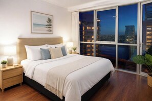 Ein modernes Schlafzimmer mit einem großen Bett, Nachttischen, Blick auf die beleuchtete Stadt und einem gerahmten Bild an der Wand.
