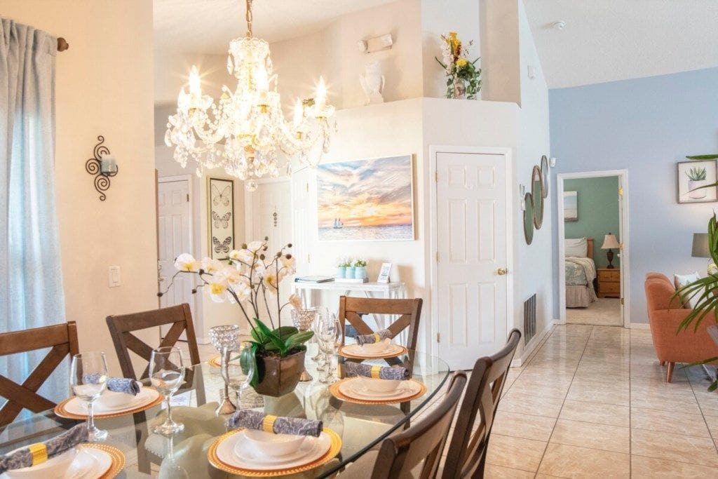 A dining area with a glass table, chairs, and a chandelier. There is a door leading to another room.