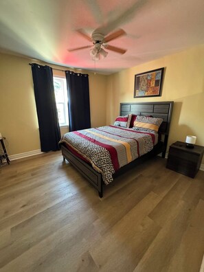 Une chambre à coucher avec un lit, un ventilateur de plafond, une table de chevet et une fenêtre avec des rideaux.