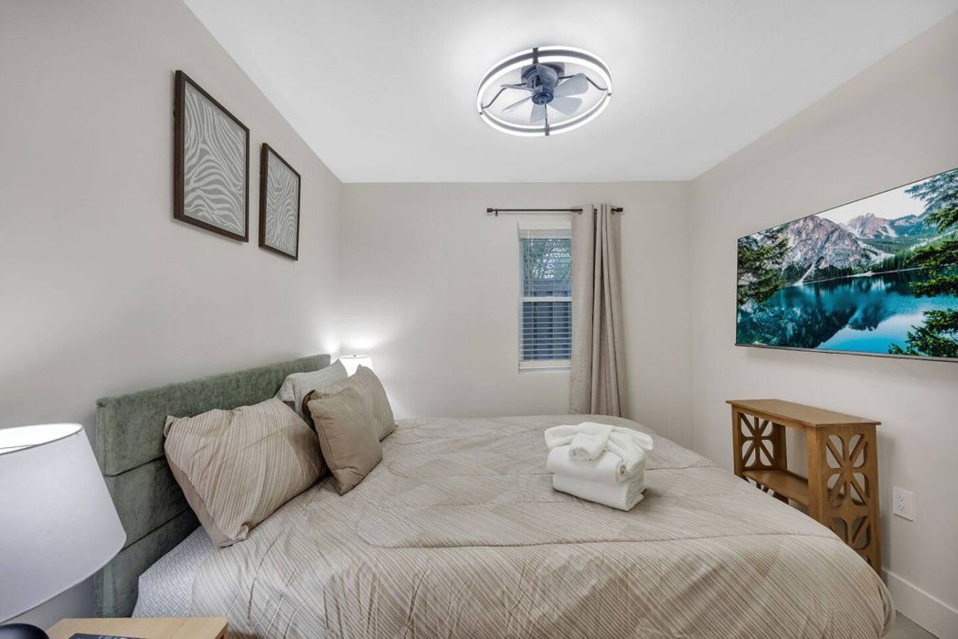 Une chambre à coucher avec un lit, une lampe de chevet, une fenêtre avec des rideaux, un ventilateur de plafond et un tableau accroché au mur.