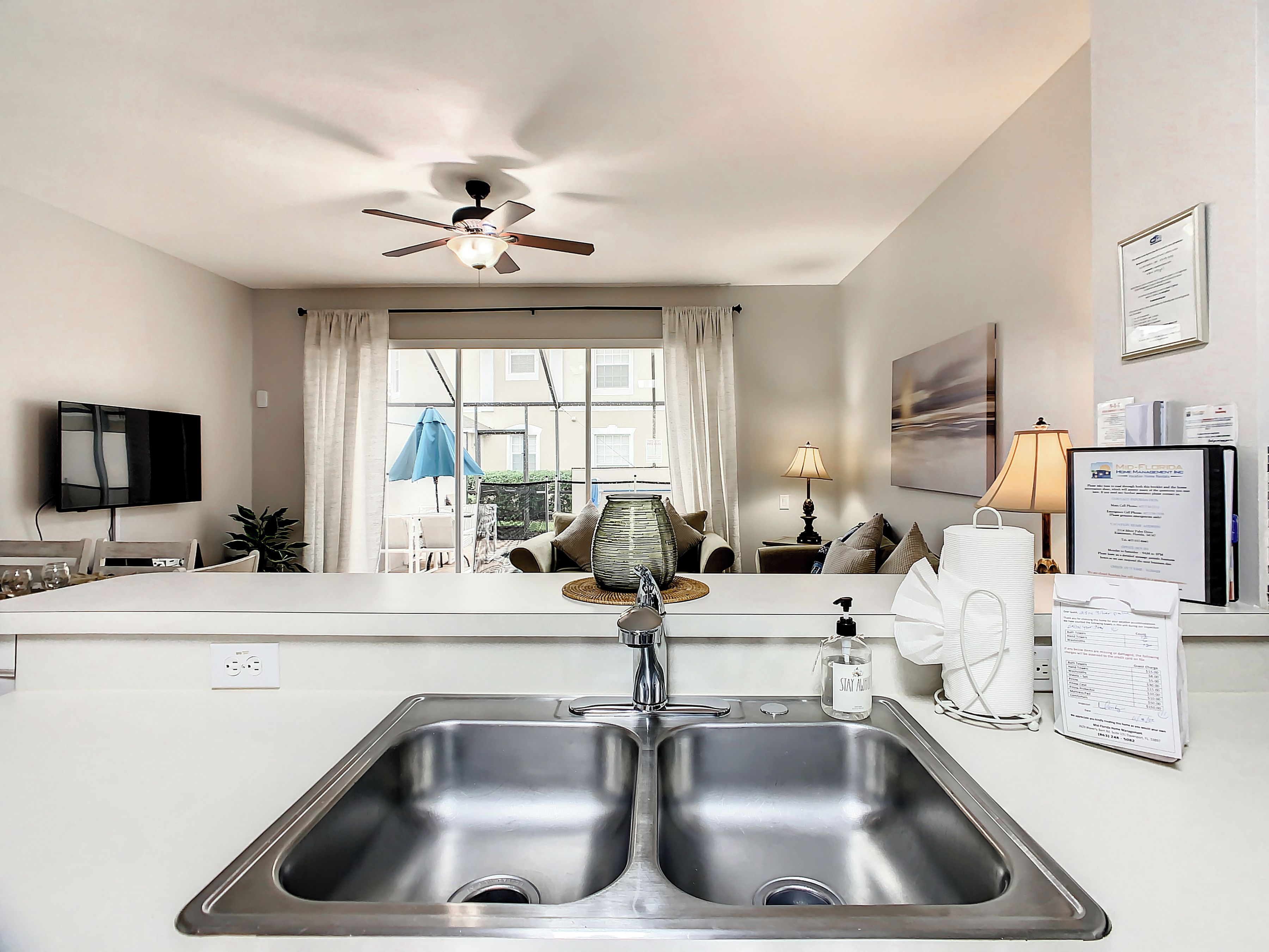 A modern kitchen with a double sink, a ceiling fan, a large window with curtains, a flat-screen TV, and a view of the outside.