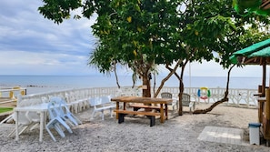 Un área de estar frente al mar con sillas de plástico y una mesa de madera, rodeada por una cerca blanca y un árbol con hojas verdes.