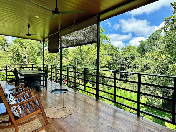 A wooden deck with a table and chairs, a small round table, and a view of lush greenery.