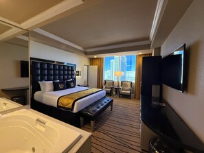 Habitación de hotel con una cama grande, un área de descanso con sillas, un televisor y vistas a la ciudad.