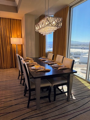 Un comedor de hotel con una mesa grande dispuesta para seis personas, una araña y vistas de la ciudad a través de amplios ventanales.