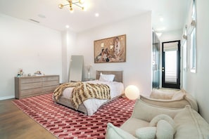 A bedroom with a bed, dresser, mirror, artwork, and a door. - Poinsettia Maison By Alora Reserve (Los Angeles)