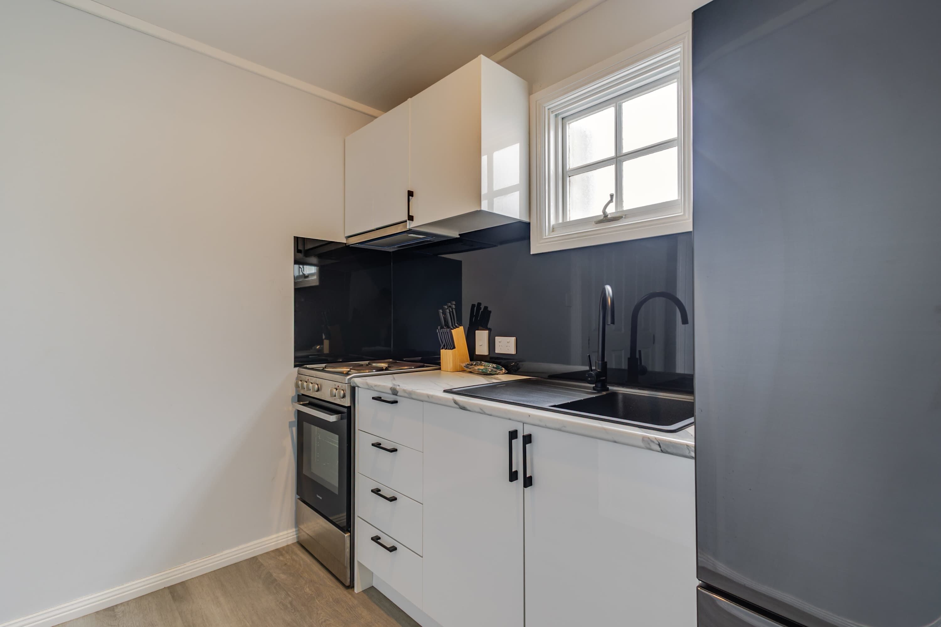 Une cuisine compacte avec des meubles blancs, une cuisinière en acier inoxydable, une hotte noire, une fenêtre au-dessus de l’évier et un robinet noir.