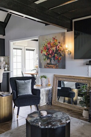 Un salon moderne avec un fauteuil noir, une table basse en marbre, un grand tableau représentant des fleurs, un miroir avec un cadre doré et une statue posée sur le sol.