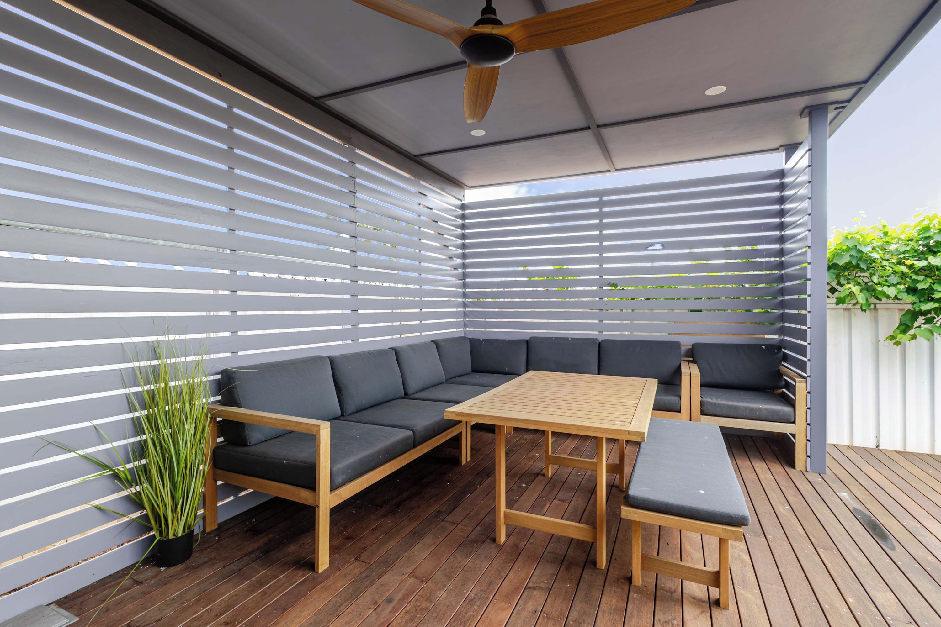 An outdoor seating area with a wooden table and benches, surrounded by a wooden deck and a slatted wall.