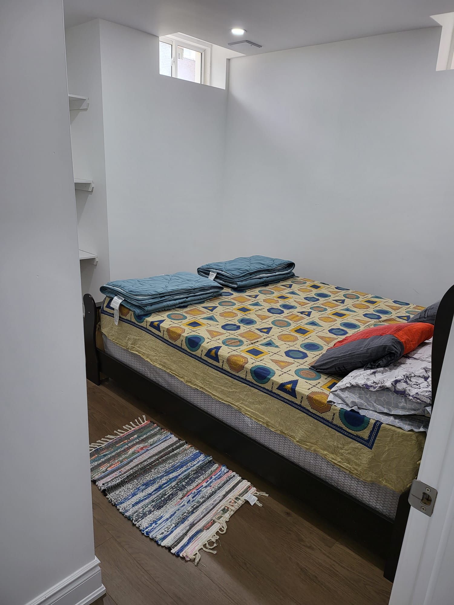 A small, single-bed room with a patterned bedspread, a striped rug, and a window with a view of a building.
