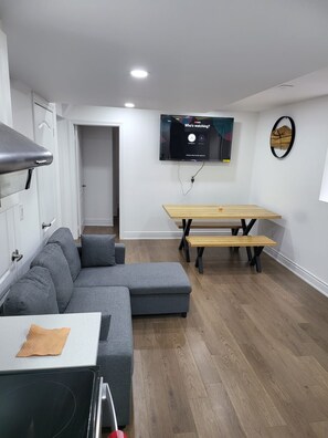 A modern living room with a grey sectional sofa, a wooden dining table with benches, a flat-screen TV mounted on the wall, and a small decorative item on the wall. - Spacious 3BR/2BA · Parking + EV · Near GO Train (Brampton)