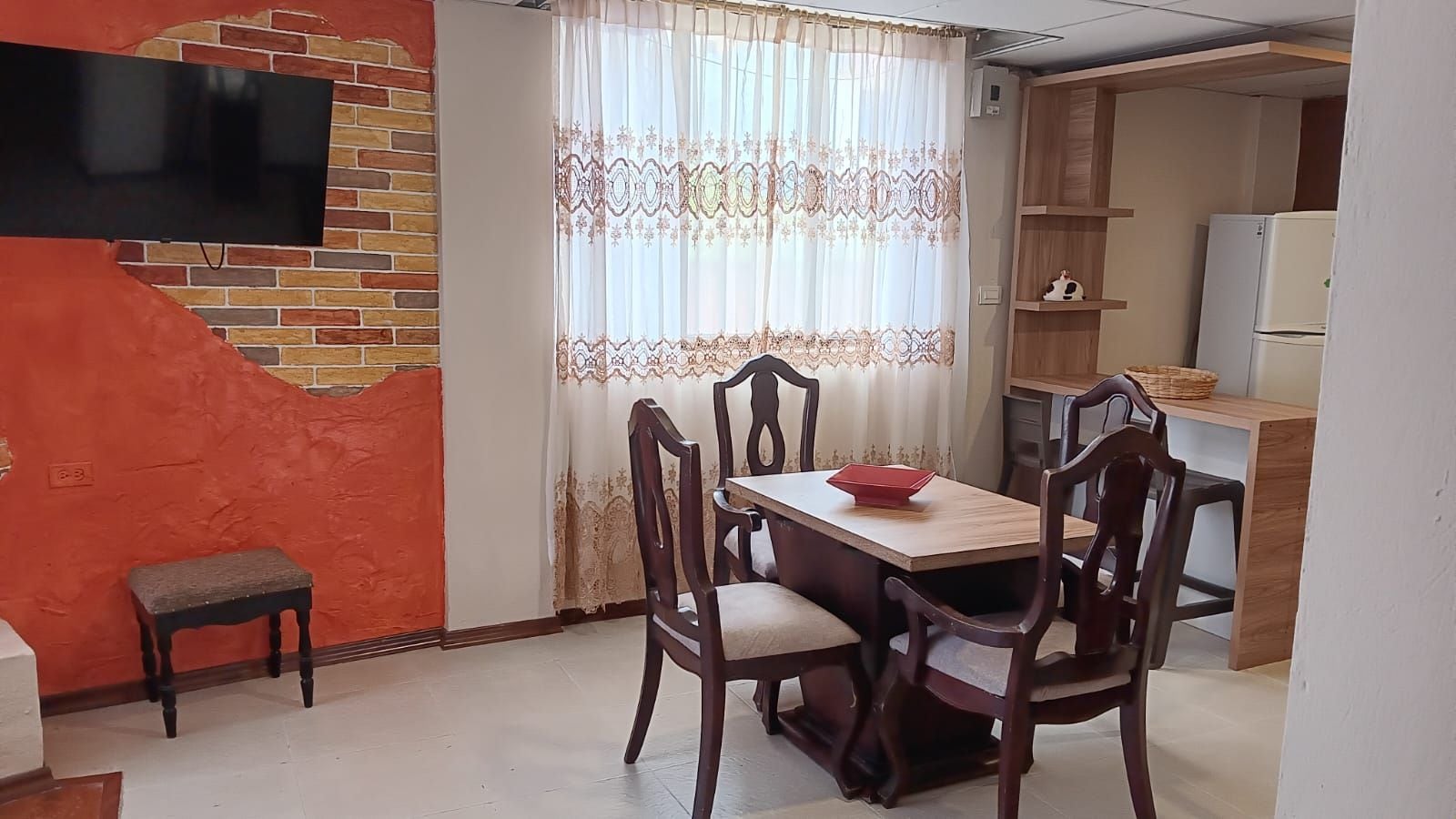 A dining area with a wooden table and chairs, a small stool, a television mounted on a brick wall, and a kitchen area with a refrigerator and shelves.