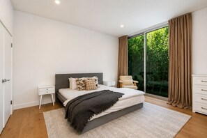 A modern bedroom with a large bed, a nightstand, a chair, and a view of greenery through a window. - Cordell Skyhouse By Alora Reserve (Los Angeles)