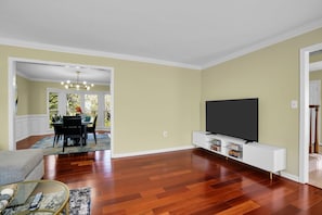 A living room with hardwood flooring, a flat-screen TV on a white stand, a dining area with a table and chairs, and a view through a window. - Elegant living at its finest in Ashburn (Ashburn)
