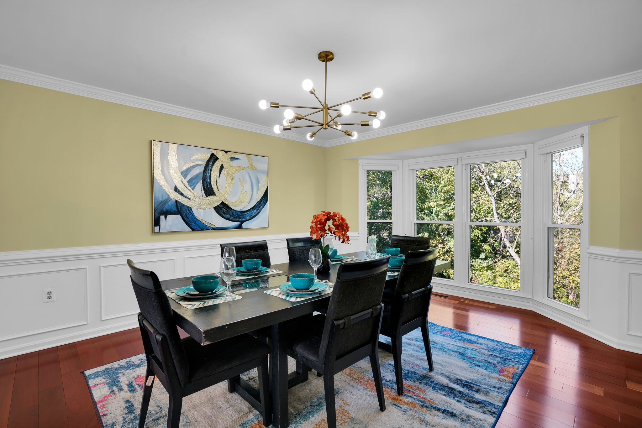 Une salle à manger avec une grande table dressée pour six personnes, entourée de chaises, un tableau au mur et une fenêtre donnant sur un paysage verdoyant.