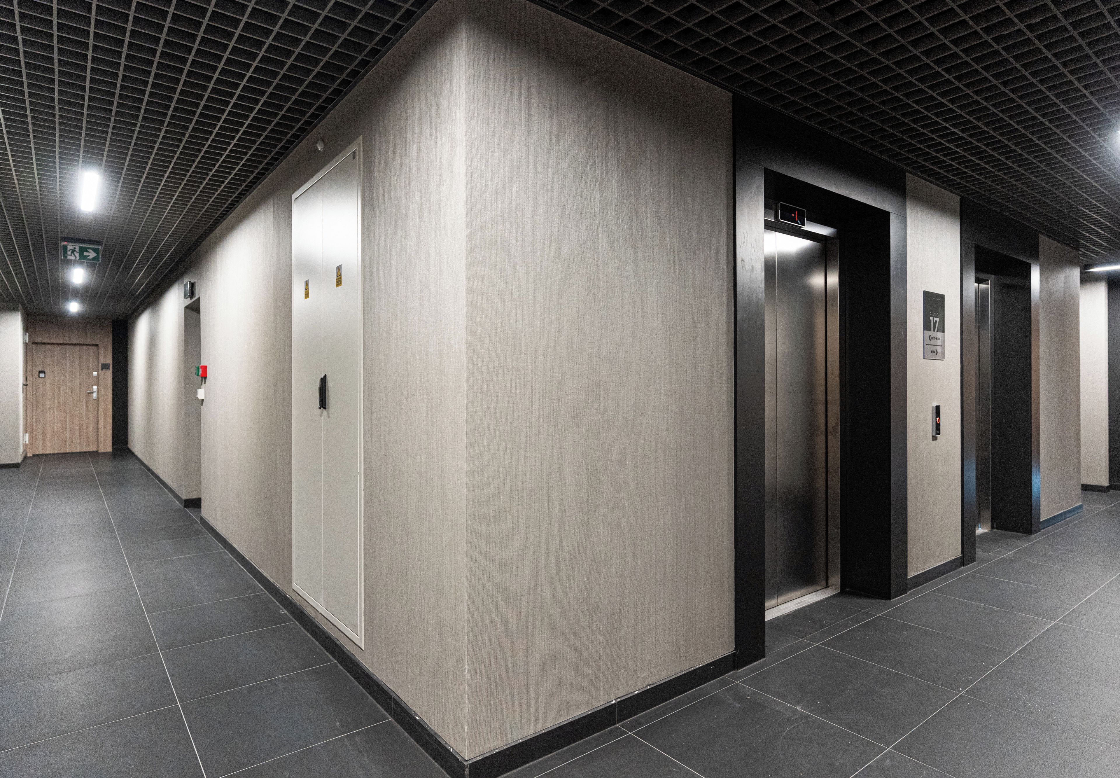 A corridor with a tiled floor, a door with an emergency exit sign, and an elevator with a digital display.