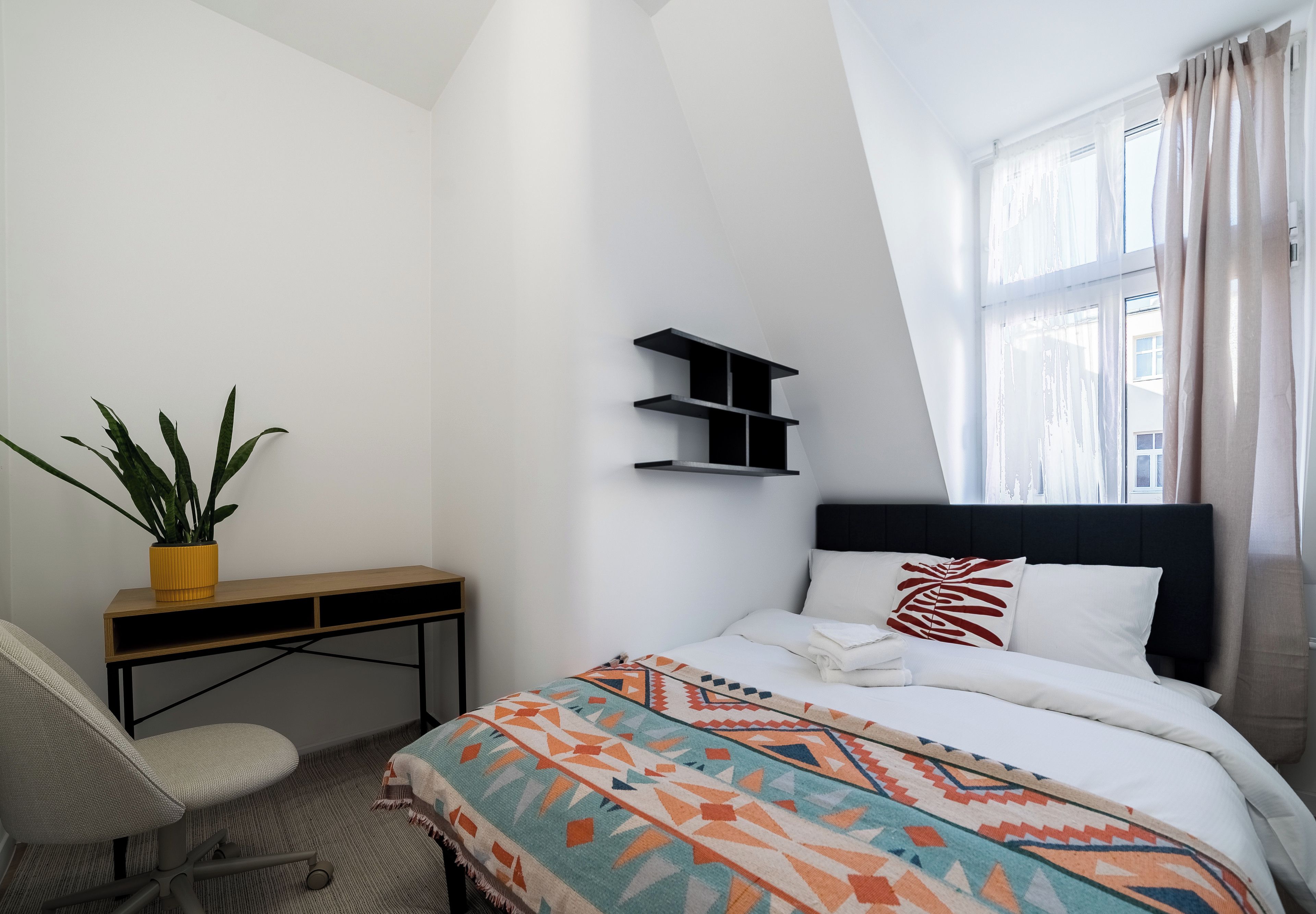 A hotel room with a bed, a desk, a chair, a potted plant, and a window with curtains.