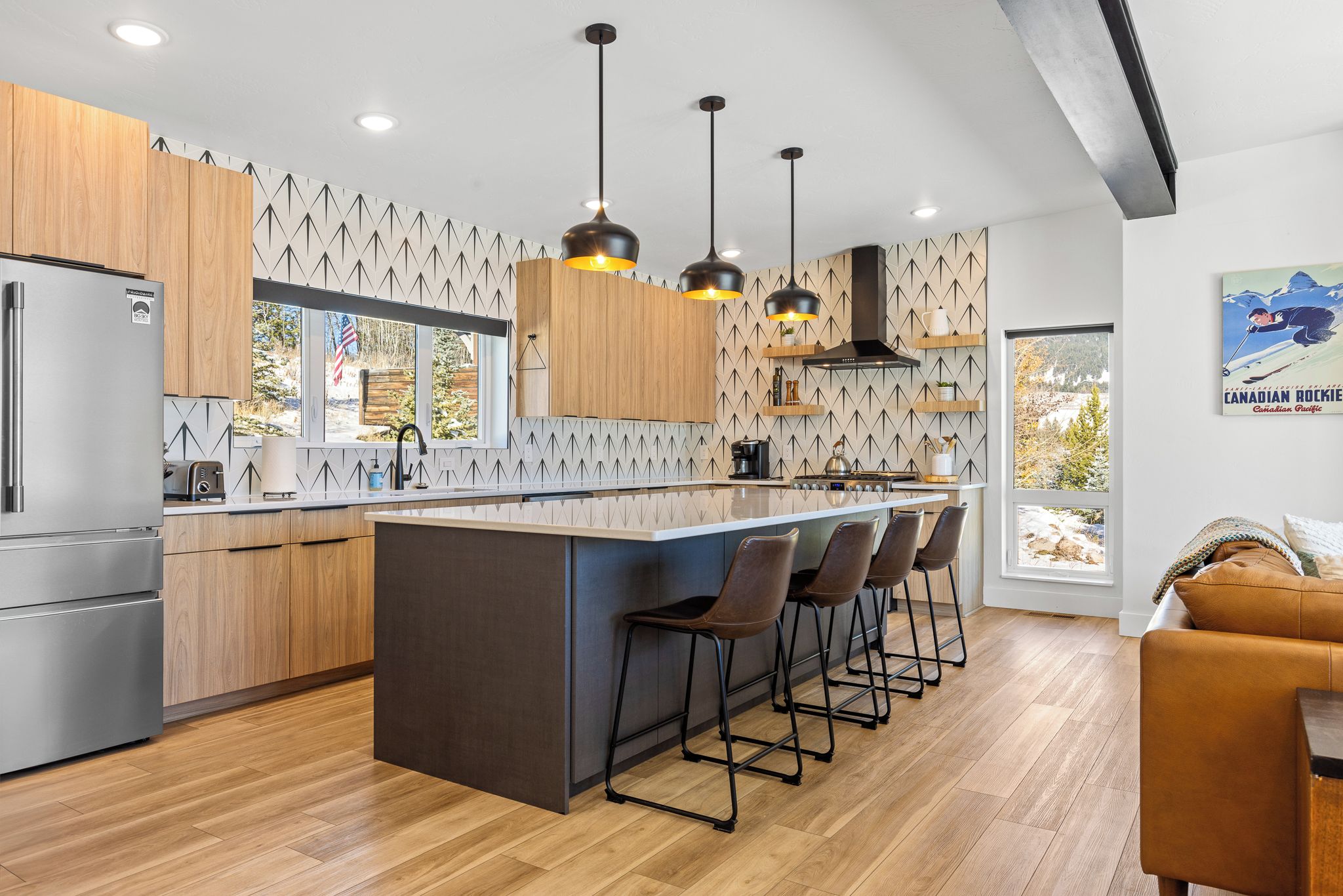 Una cucina moderna con mobili in legno, un'isola centrale con sgabelli da bar, illuminazione a sospensione e vista all'esterno attraverso ampie finestre.