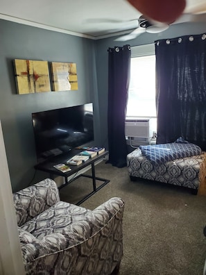 Ein Wohnzimmer mit einem Flachbildfernseher, zwei gemusterten Sofas, einem Couchtisch mit Büchern und einem fenster mit Vorhängen.