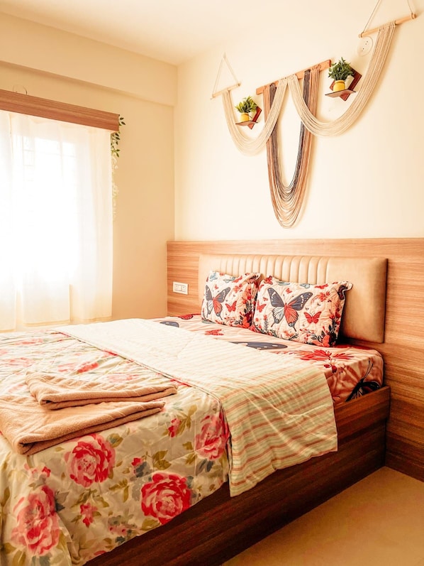 Un dormitorio con una cama, ropa de cama floral, cabecera de madera y un colgador de macramé con plantas.