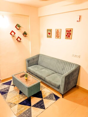 Una sala de estar con un sofá gris, una mesa de centro y un tapete estampado. Se observan decoraciones colgantes en la pared y una puerta con diseño geométrico.