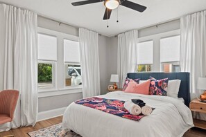 A bedroom with a bed, a chair, a nightstand, a lamp, a ceiling fan, and a window with curtains. - Tampa Oasis - Private Heated Pool, Seminole Heights (Tampa)