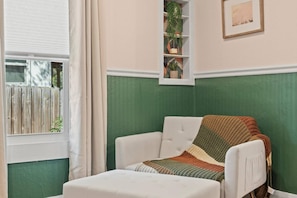A room with a green accent wall, a white armchair, a patterned throw blanket, a small shelf with plants, and a window with a view of a wooden fence. - Tampa Oasis - Private Heated Pool, Seminole Heights (Tampa)