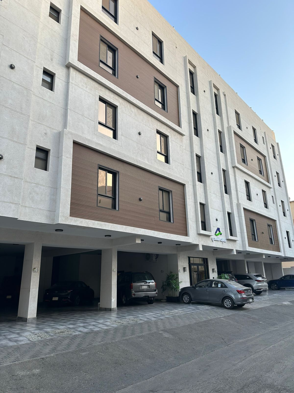 Un edificio de varios pisos con un diseño arquitectónico moderno, que presenta una combinación de fachadas de ladrillo y concreto, y un área de estacionamiento con varios automóviles.