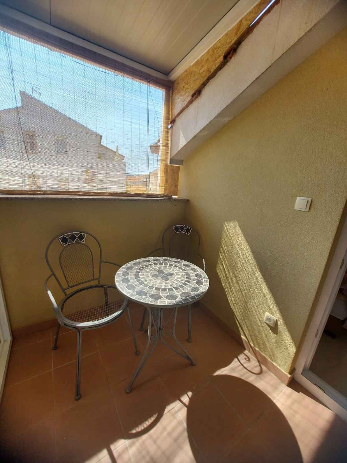 Un pequeño balcón con dos sillas de metal y una mesa redonda, una ventana con persianas, y vista a edificios.