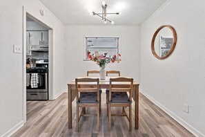 A dining area with a wooden table and chairs, a vase with flowers, a window with blinds, and a kitchen visible through an open door. - Cozy Montrose Retreat, King Bed + Parking (Houston)