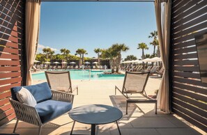 A poolside area with lounge chairs and a small table, overlooking a swimming pool with umbrellas and palm trees. - Beach House | Resort King Room | Outdoor Pool (Hilton Head Island)