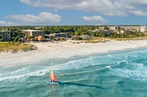 En badehotel med sejlbåd på vandet, villaer direkte ved stranden og en klar himmel.