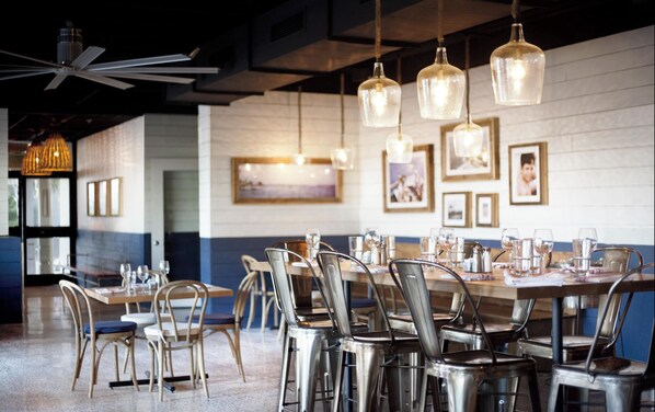 En restaurantindretning med høje borde, metalstole, hængende pendellamper og billeder i rammer på væggen.