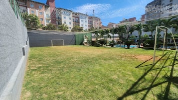 Un área al aire libre cercada con un campo de césped, un arco de fútbol y una piscina.