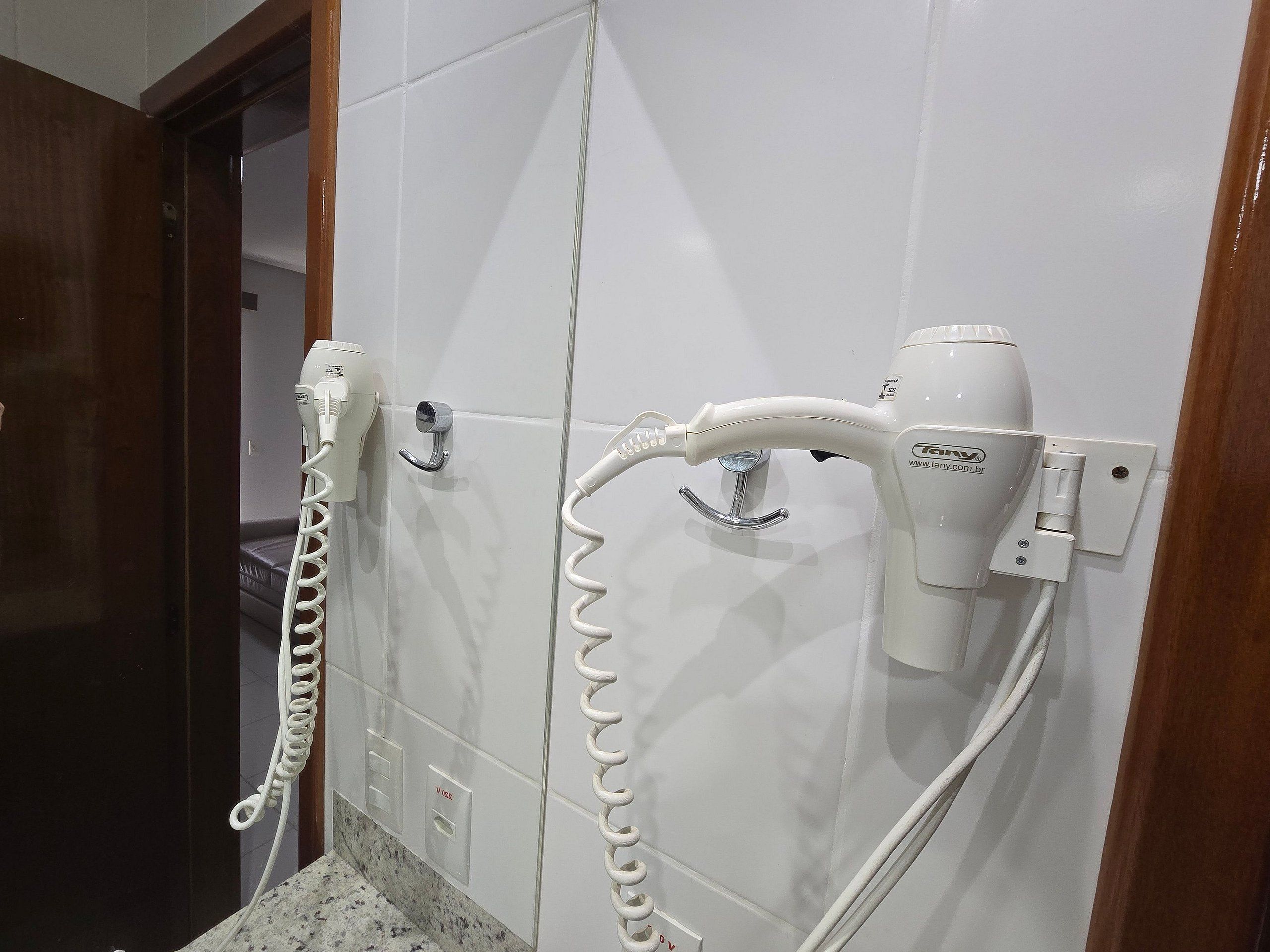 Un baño con paredes de azulejos blancos, una encimera de mármol y dos secadores de pelo montados en la pared.