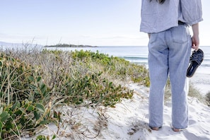 A person standing on a sandy beach with vegetation, holding a pair of shoes, and facing the ocean. - Oystercatcher Bicheno - Private track to the beach! (Glamorgan-Spring Bay)