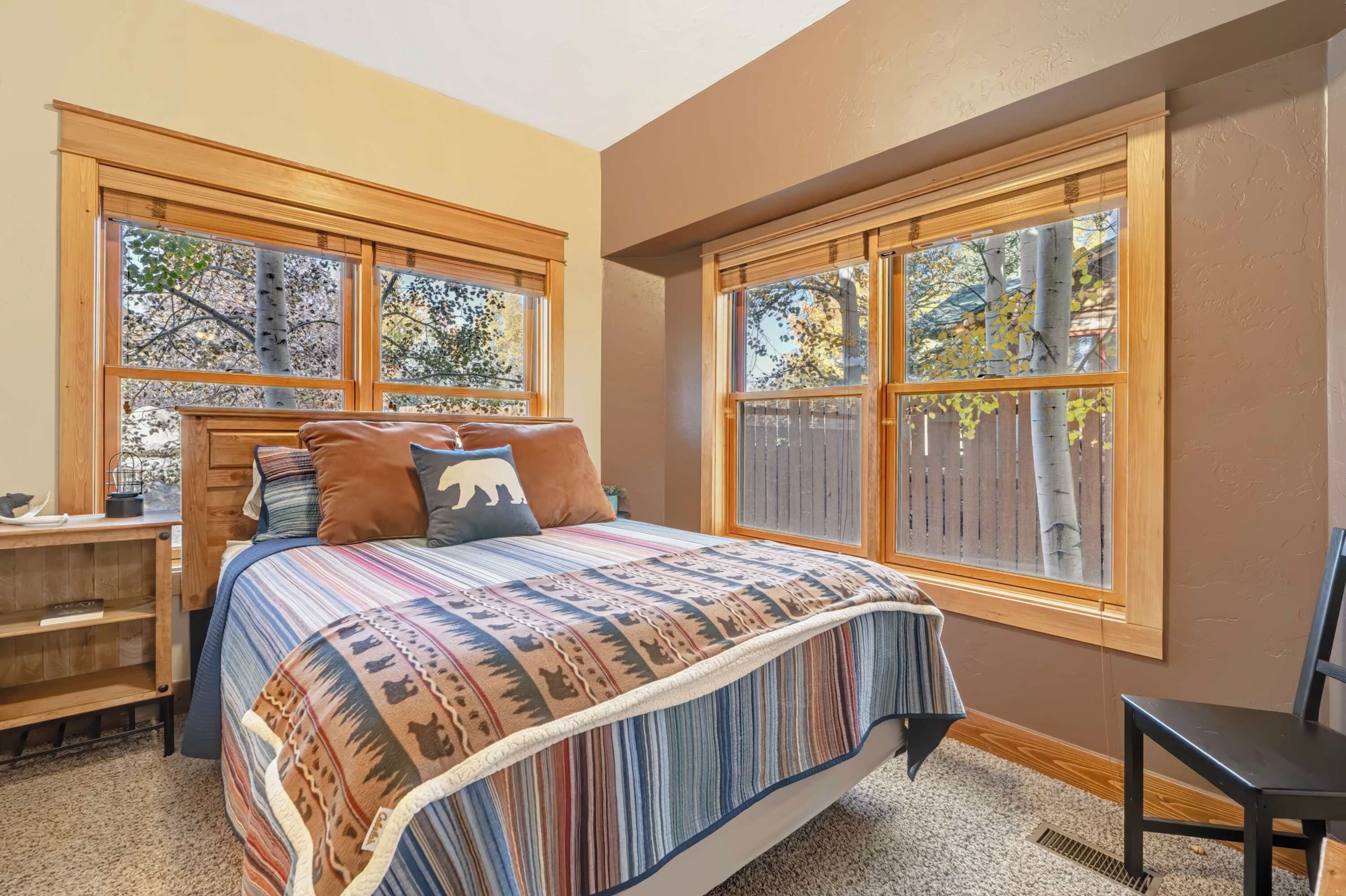 A bedroom with a large bed, a wooden nightstand, and a chair. There are two windows with a view of trees.