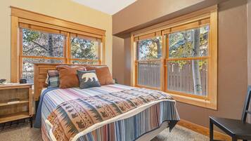 A bedroom with a large bed, a wooden nightstand, and a chair. There are two windows with a view of trees.
