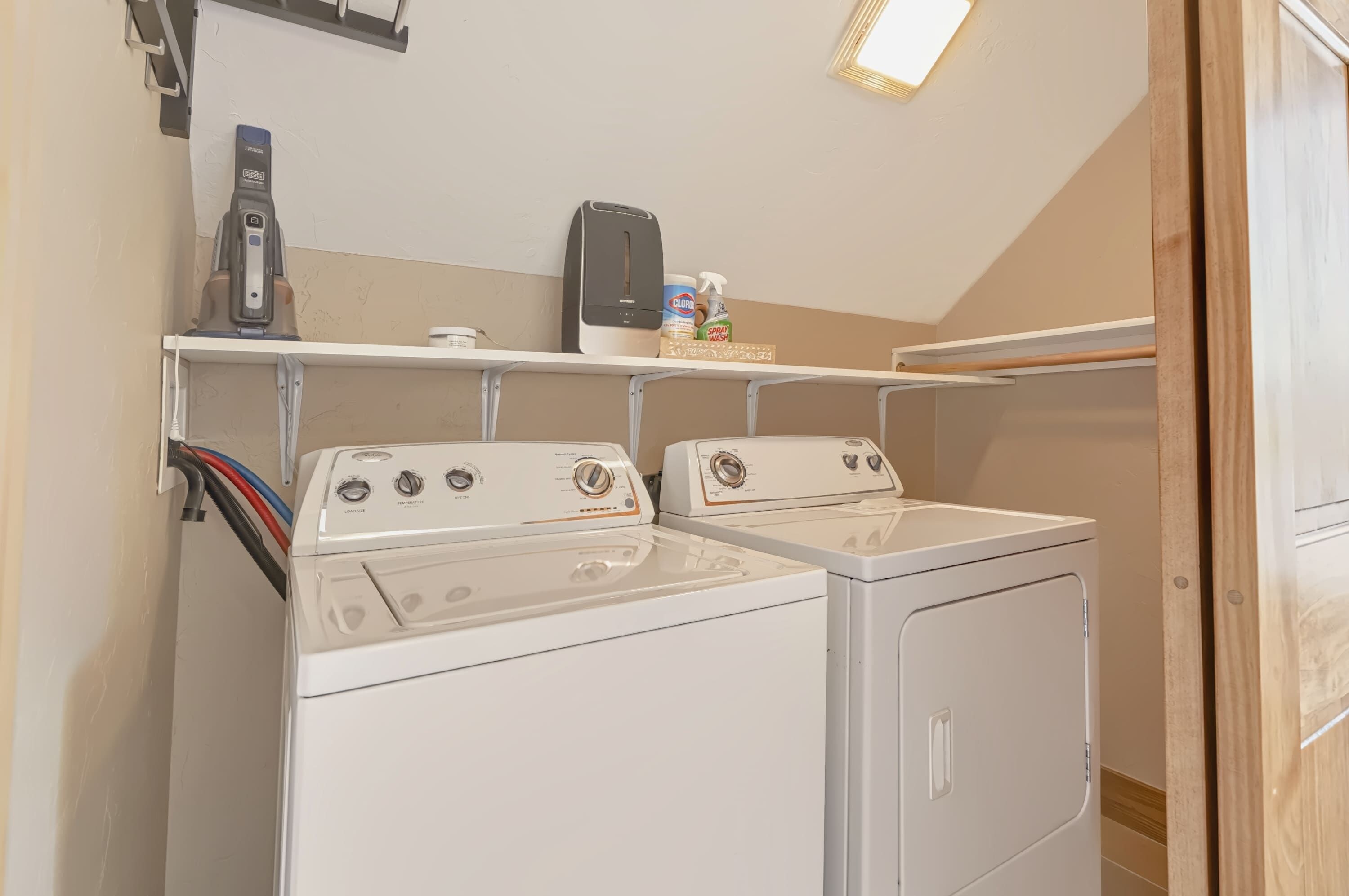 A laundry area with two front-loading washers and dryers, a shelf with cleaning supplies, and a ceiling light fixture.