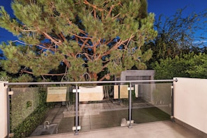 Ein moderner Balkon mit Glasgeländer, einem Metallgitter und einem Baum im Hintergrund.