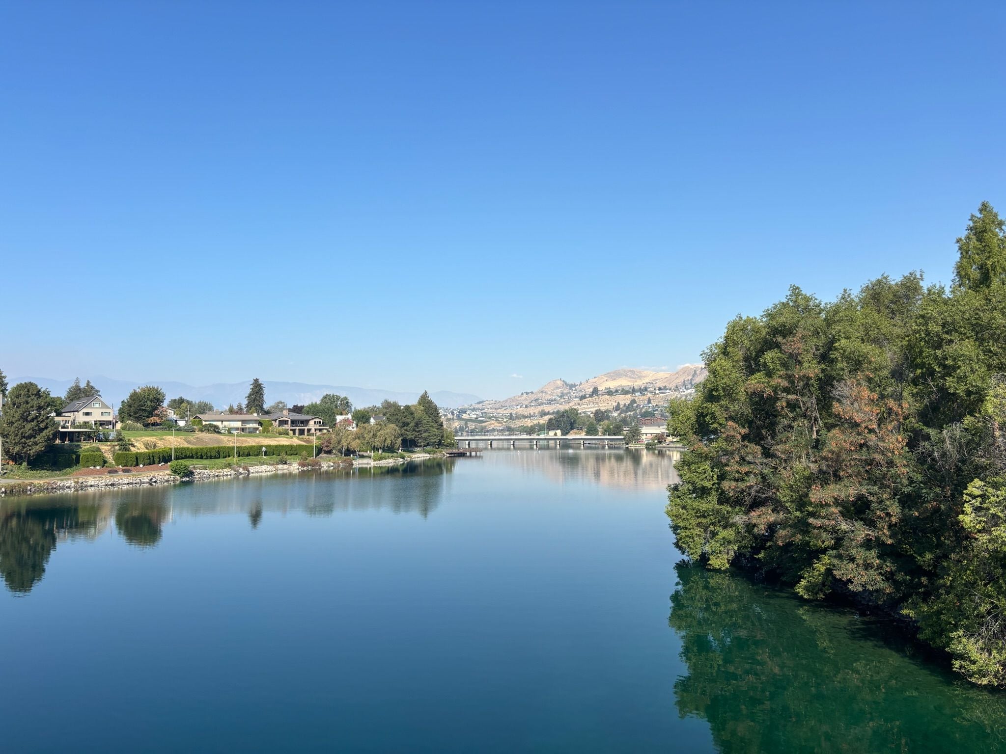 A calm lake with a bridge, surrounded by trees and houses.