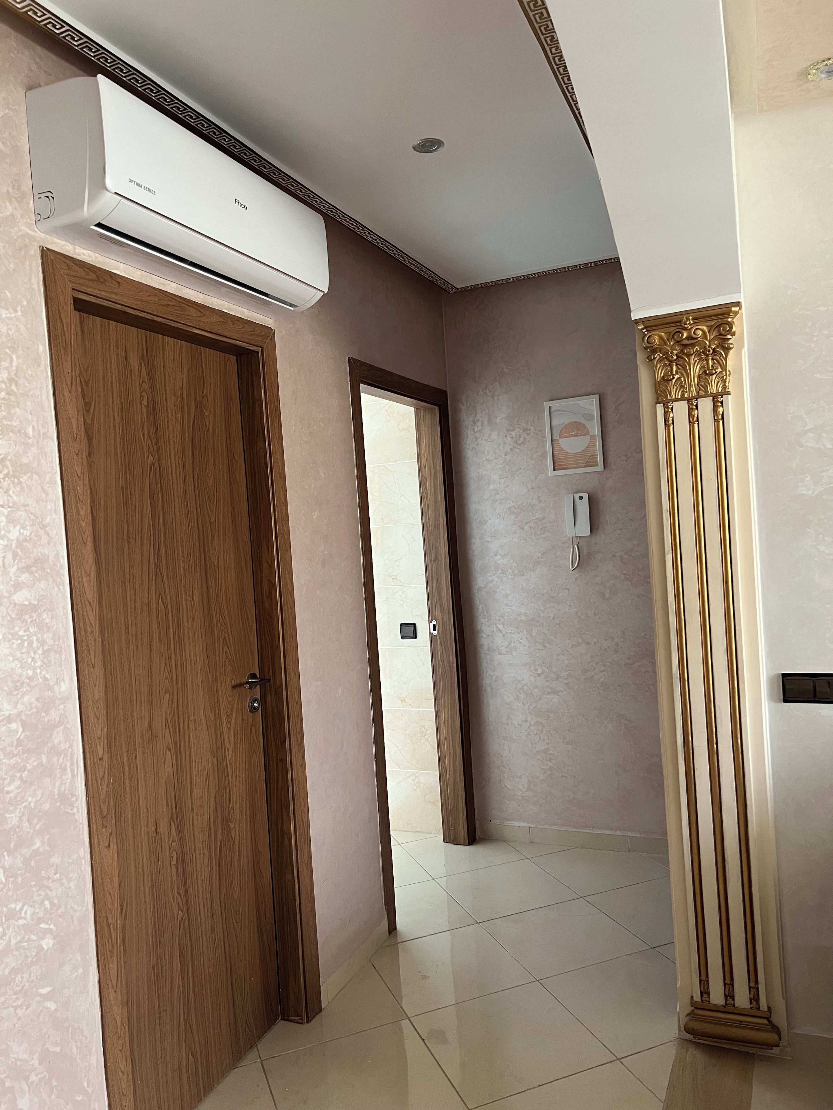 Un couloir avec une porte en bois, un climatiseur blanc, un sol carrelé et une colonne décorative.