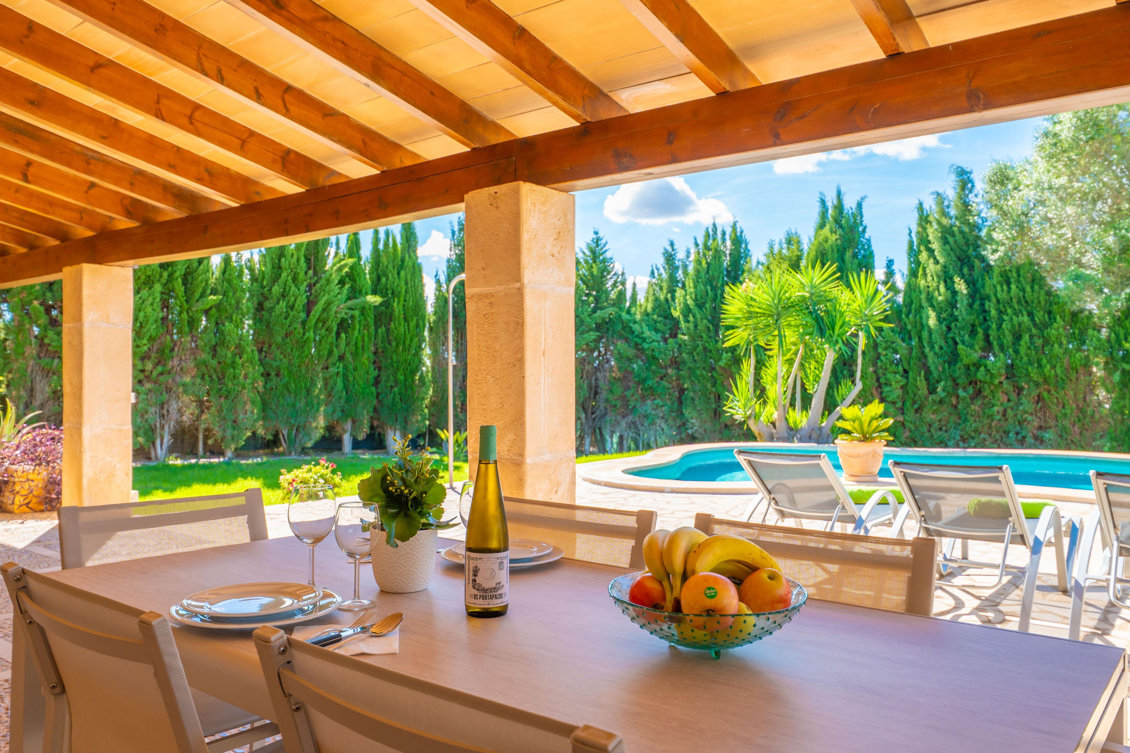 An outdoor dining area with a pool view, wooden ceiling, and a table set with a wine bottle and fruit bowl.